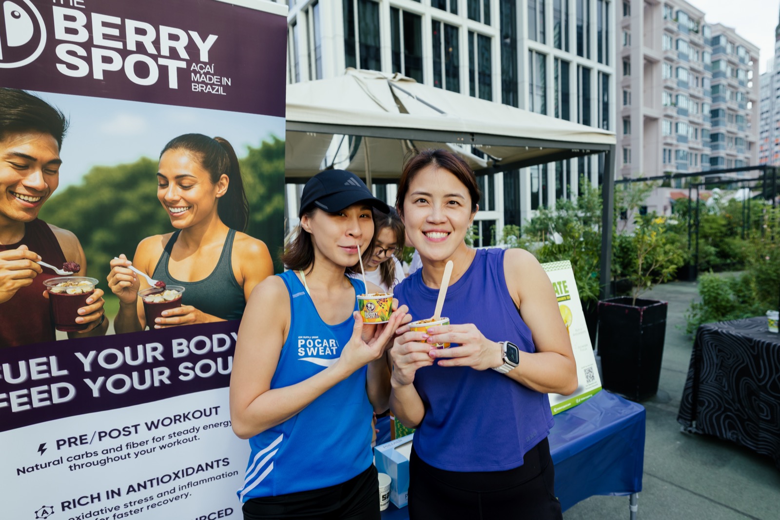 Two women holding açaí cups with wooden spoons in front of The Berry Spot banner
