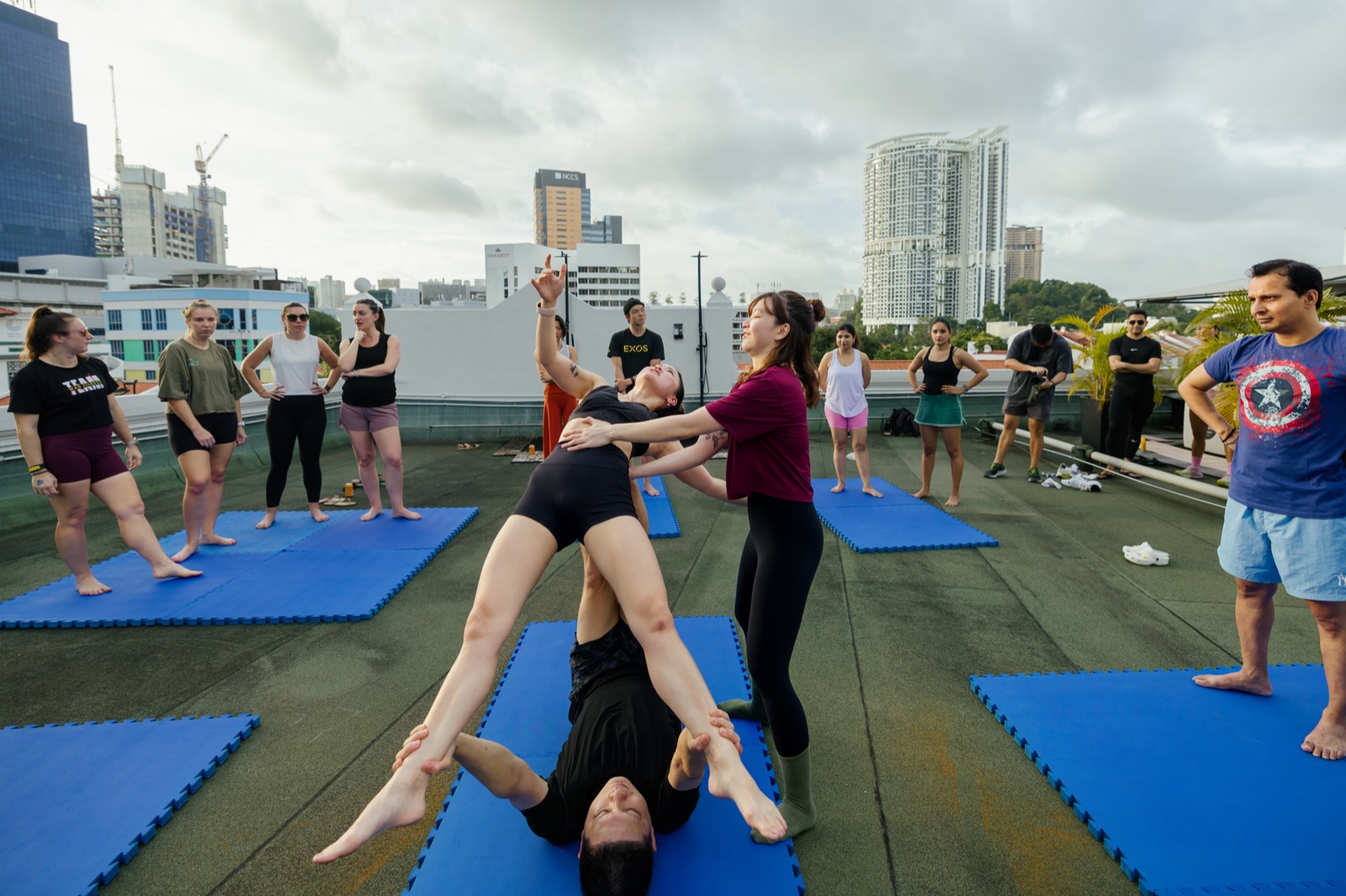 Partner acrobatics on a mat: one person balanced face-down on another's raised feet while a third spots them, the rest of the class watching