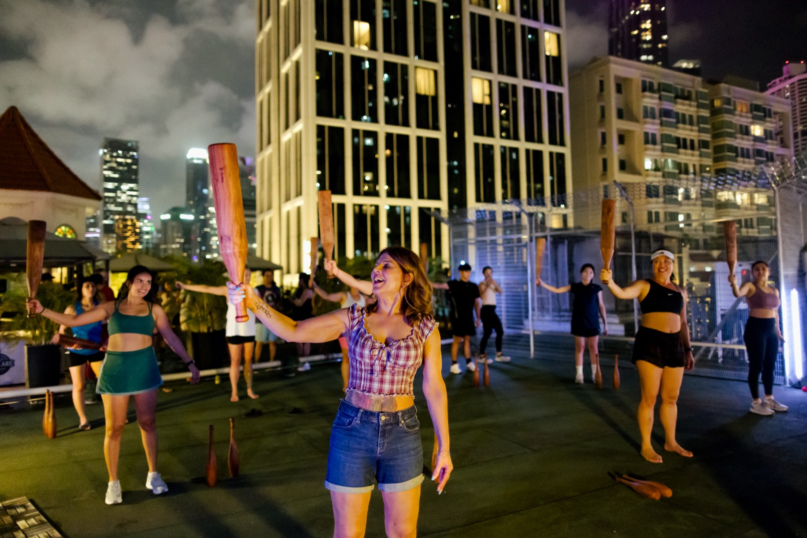ClubBells class after dark, wooden clubs raised overhead, lit apartment towers behind the rooftop