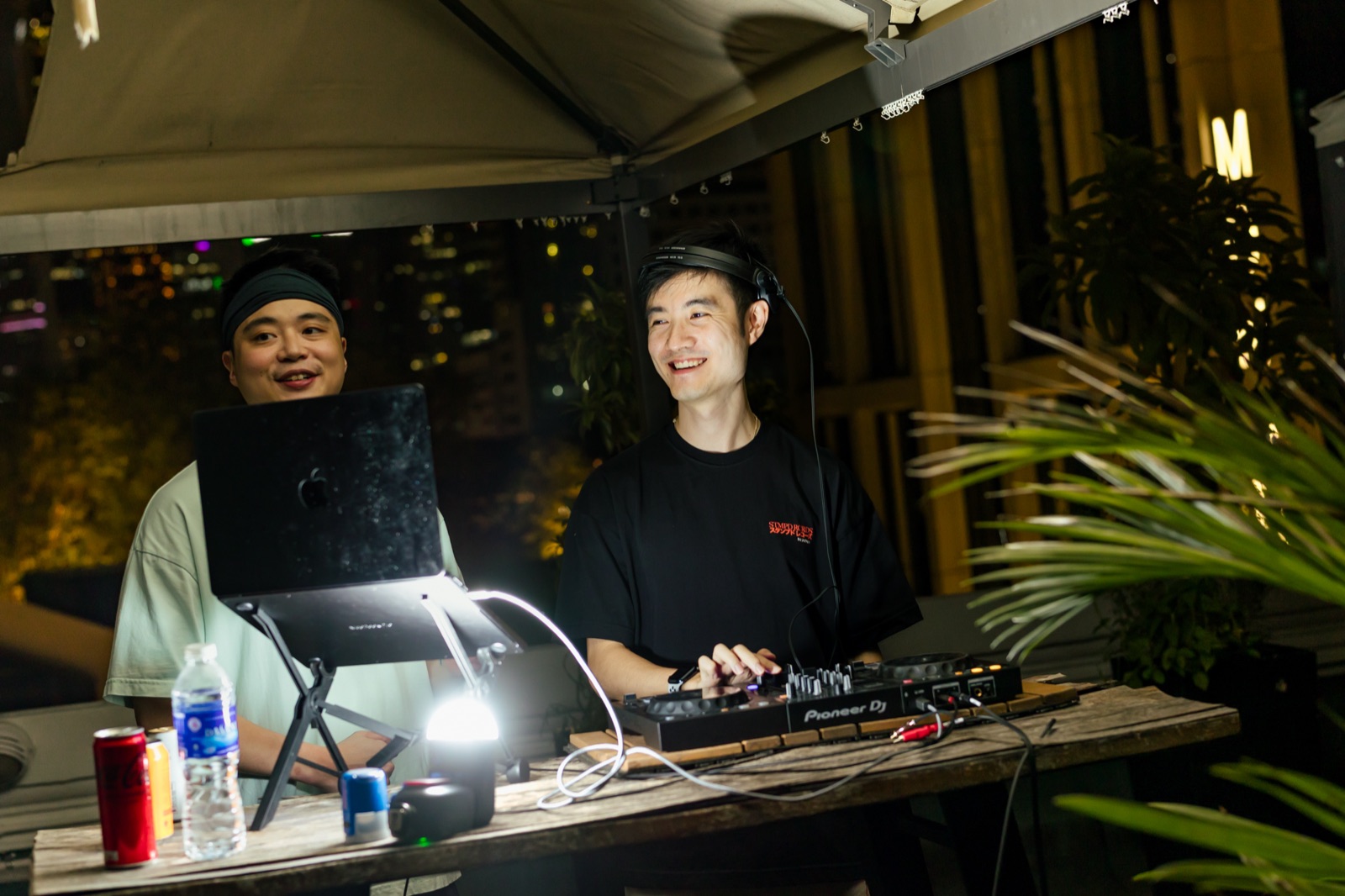 Two DJs behind a controller and a laptop under the canopy at night, city lights out of focus behind them