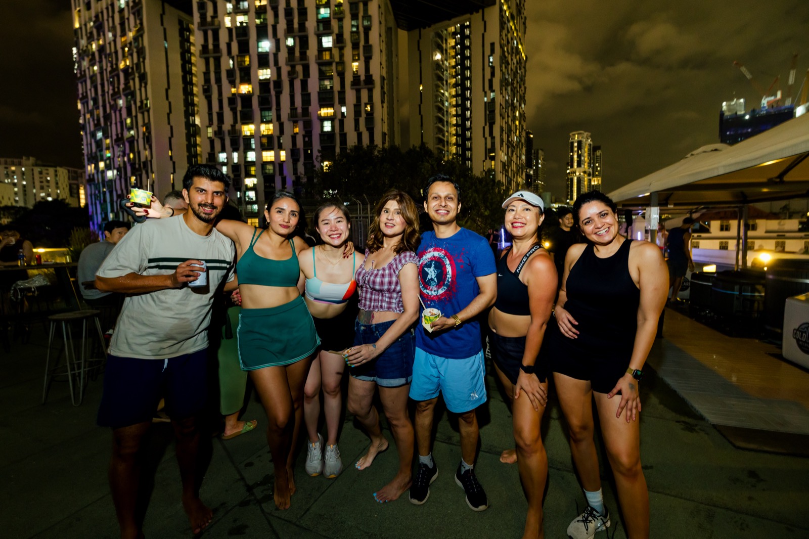 Eight people standing shoulder to shoulder for a photo at night, drinks in hand, lit apartment blocks behind