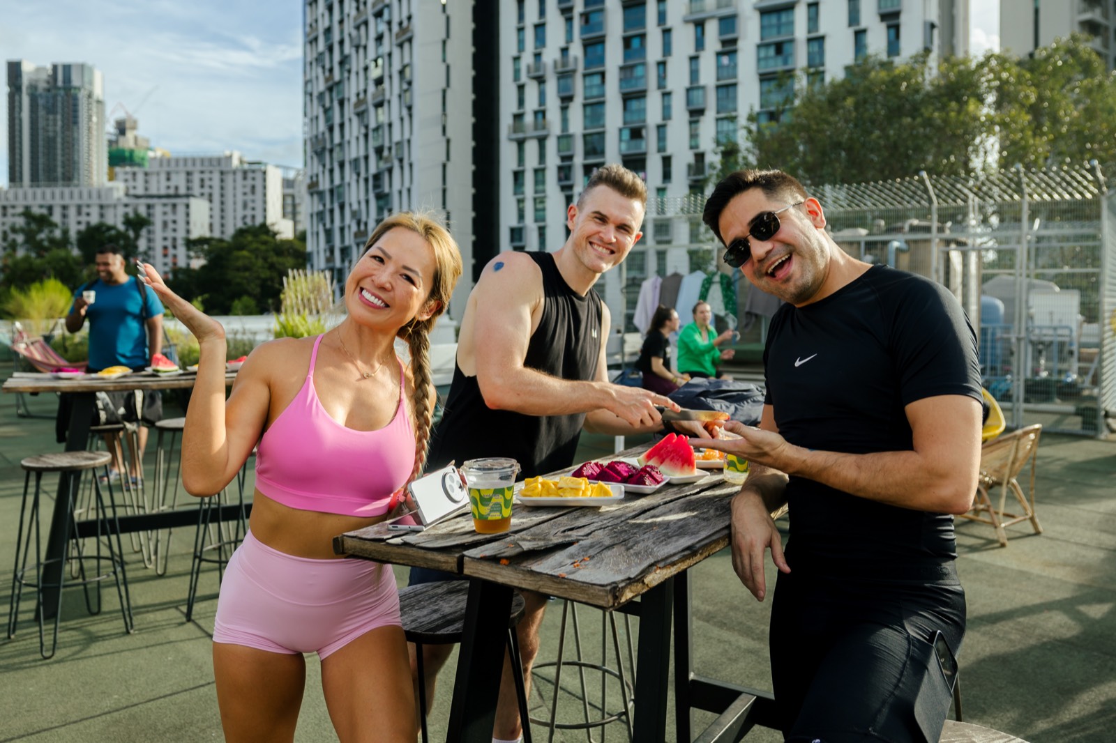 Three people laughing around a rooftop table of cut fruit: watermelon, dragonfruit and pineapple on plates, high-rise towers behind