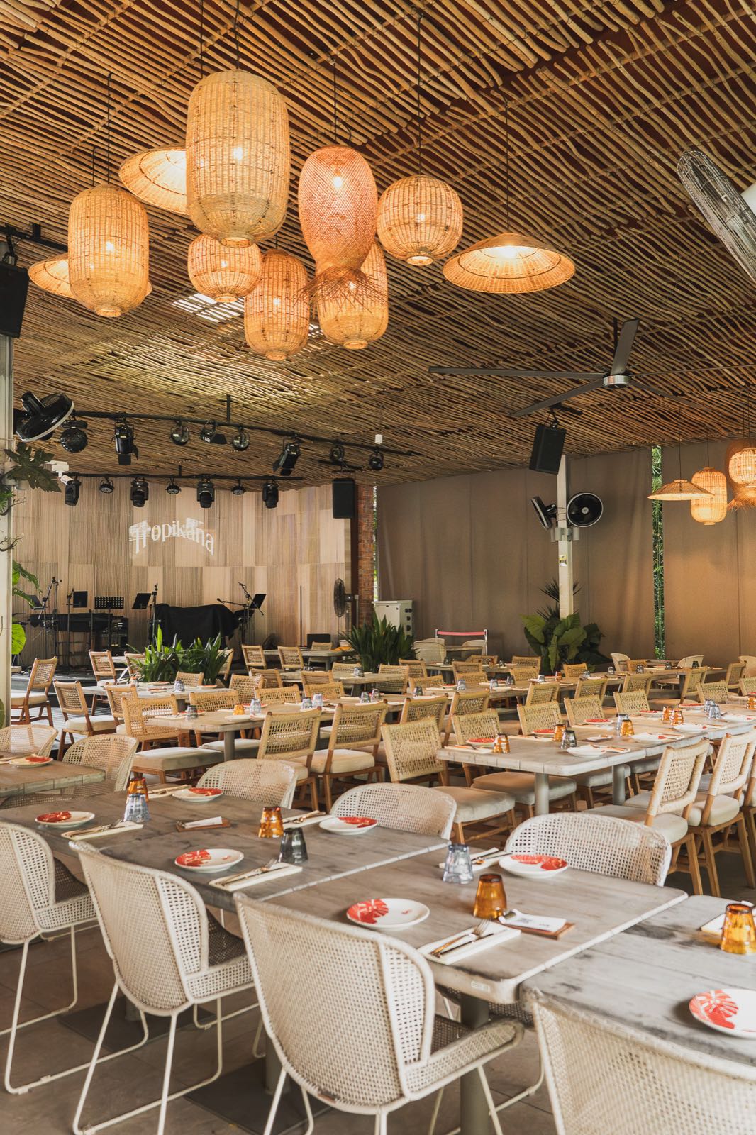 A covered dining room under a bamboo ceiling hung with woven rattan lanterns, long tables laid with plates and glasses, a stage and the Tropikãna sign at the far end