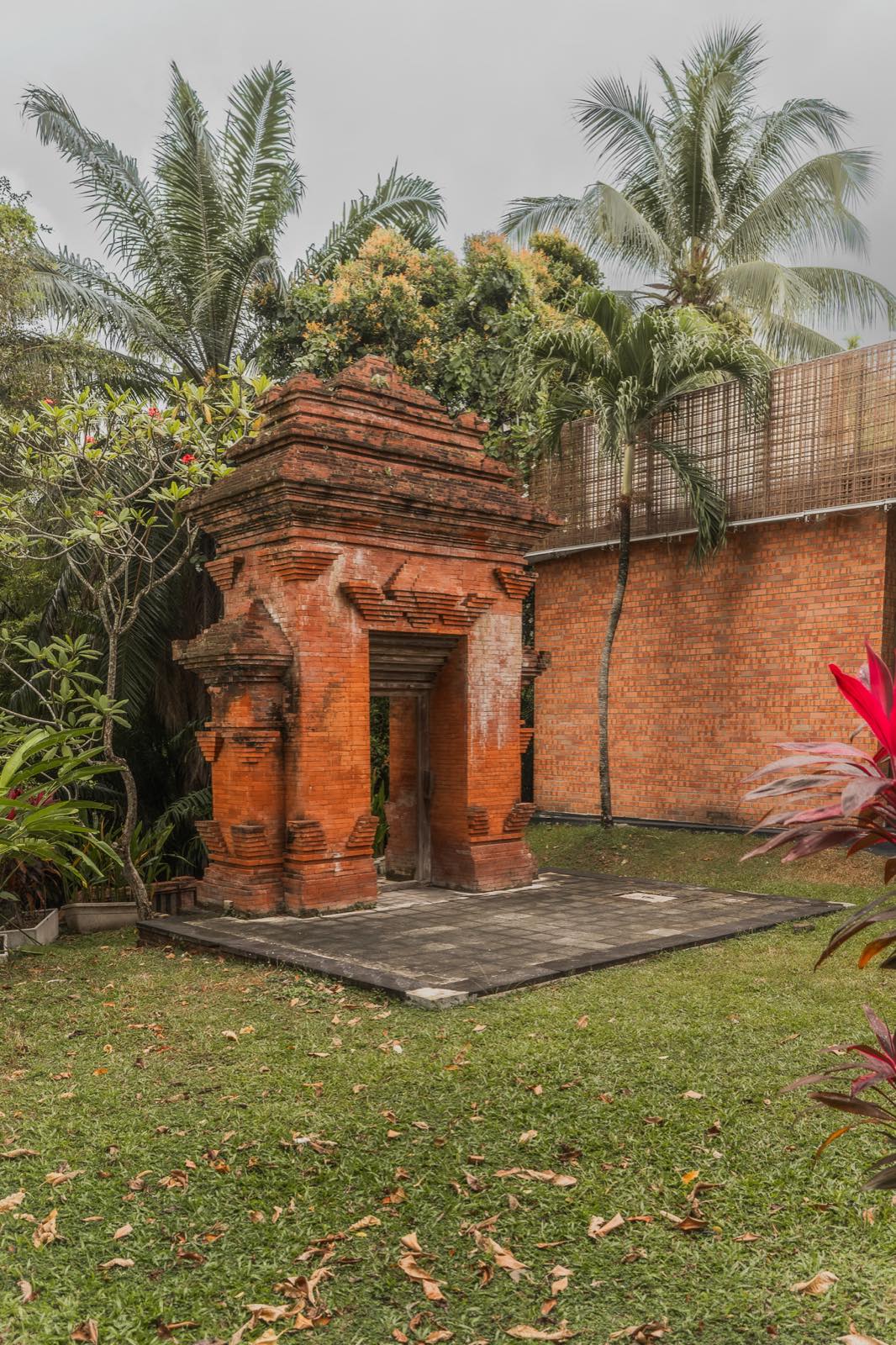 A red brick arch on a paved square in the middle of a lawn, palms, a frangipani tree and a brick wall behind it